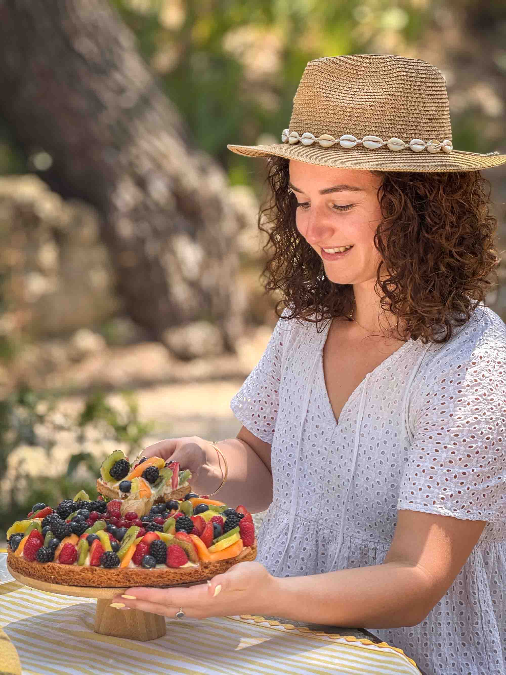 Savouring the treasures of the Provençal terroir in the heart of nature A smiling young woman wearing a straw hat decorated with shells holds a beautiful tart topped with red and exotic fruits.