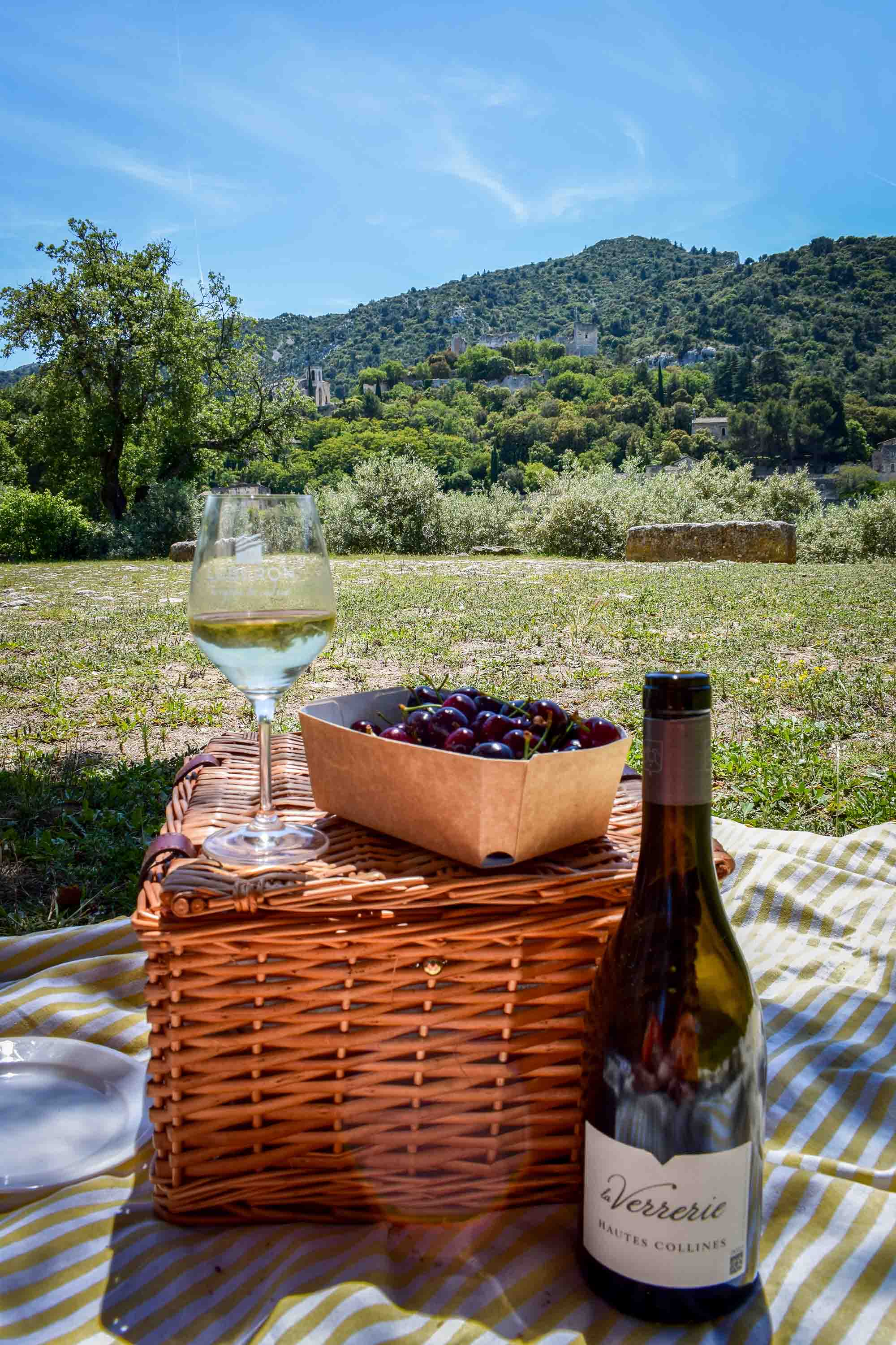 The gentle art of living and gourmet pleasures in the heart of old Oppède Picnic composition: a bottle of Verrerie wine and a filled glass placed next to a closed wicker basket topped with a punnet of fresh black cherries, all set on a striped blanket facing the Oppède landscape.