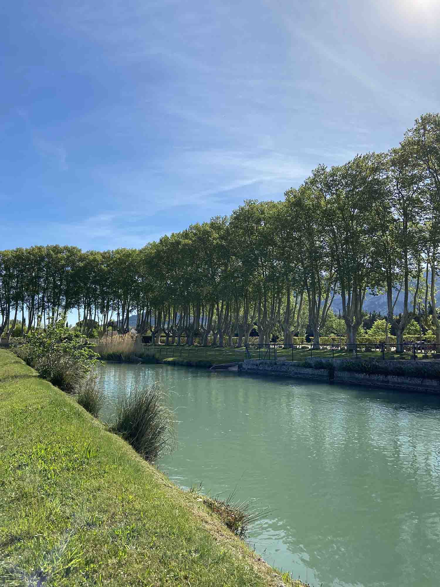 A refreshing break by the water at the Moulin Saint-Pierre View of the Moulin Saint-Pierre in Les Taillades, with its large wooden paddle wheel beside the canal, surrounded by greenery and flowers under a blue sky.